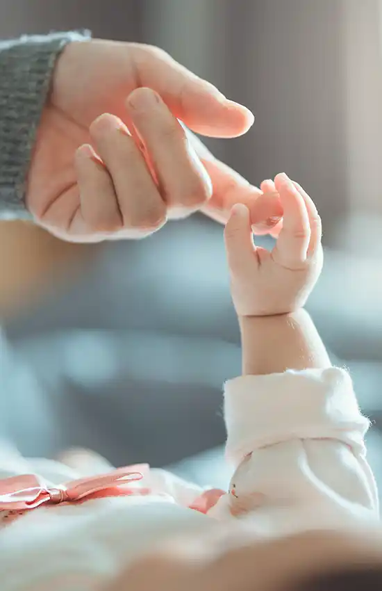 craniosacrale-therapie-babys-kinder-mobil Eine erwachsene Hand berührt behutsam die kleine Hand eines Babys, das im hellen Licht liegt und einen weichen Stoffärmel trägt.