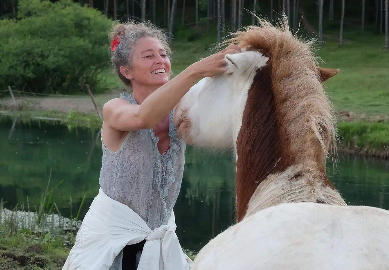 craniosacrale-therapie-pferd-mobil Eine lächelnde Frau berührt ein hellbraun-weißes Pferd am Kopf bei der Cranio-Sacrale Therapie am Ufer eines stillen Sees vor grünem Wald.