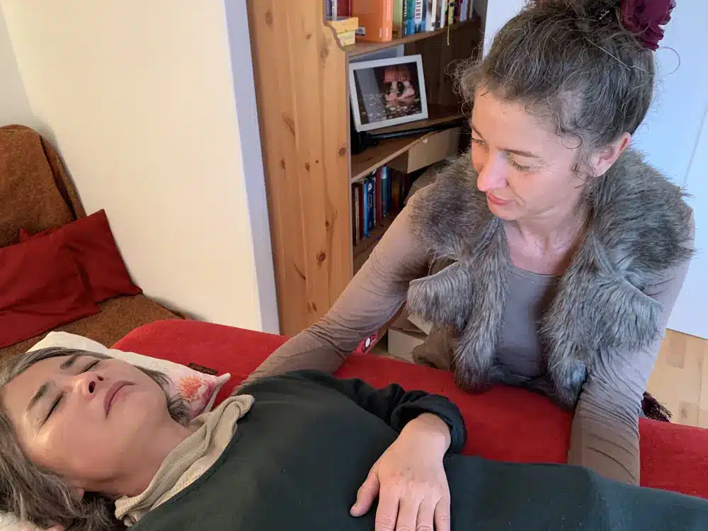 Alma legt einer liegenden Patientin behutsam die Hand auf die Schulter.