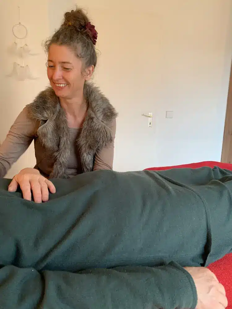 Alma sitzt lächelnd an der Liege, die Hände ruhen auf einer Patientin.