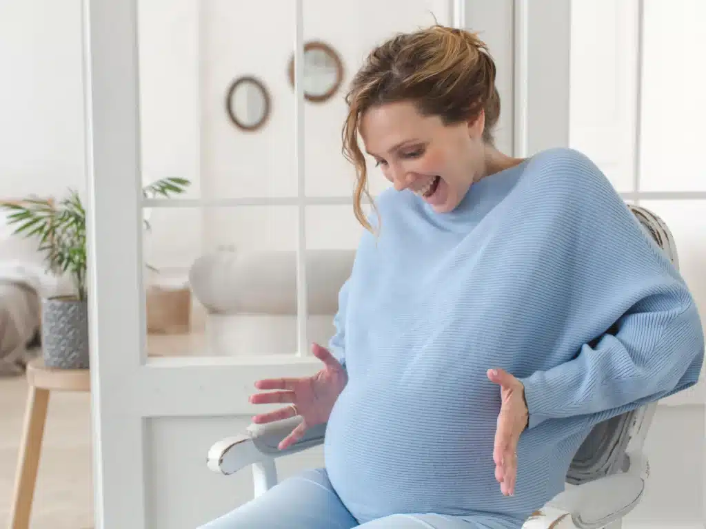 Eine schwangere Frau im hellblauen Pullover sitzt lachend auf einem Stuhl.